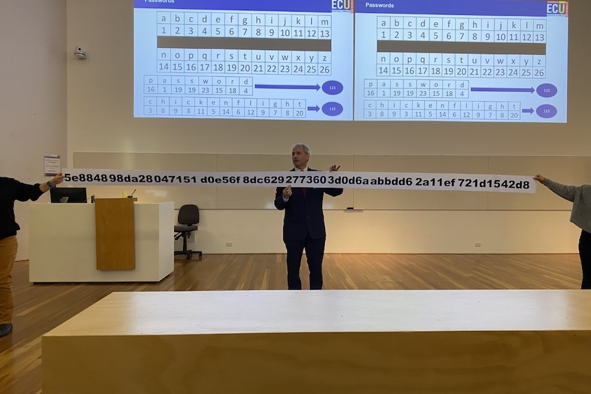 A man in a suit giving a lecture is holding a ridiculously long piece of paper with a password of random letters and numbers.