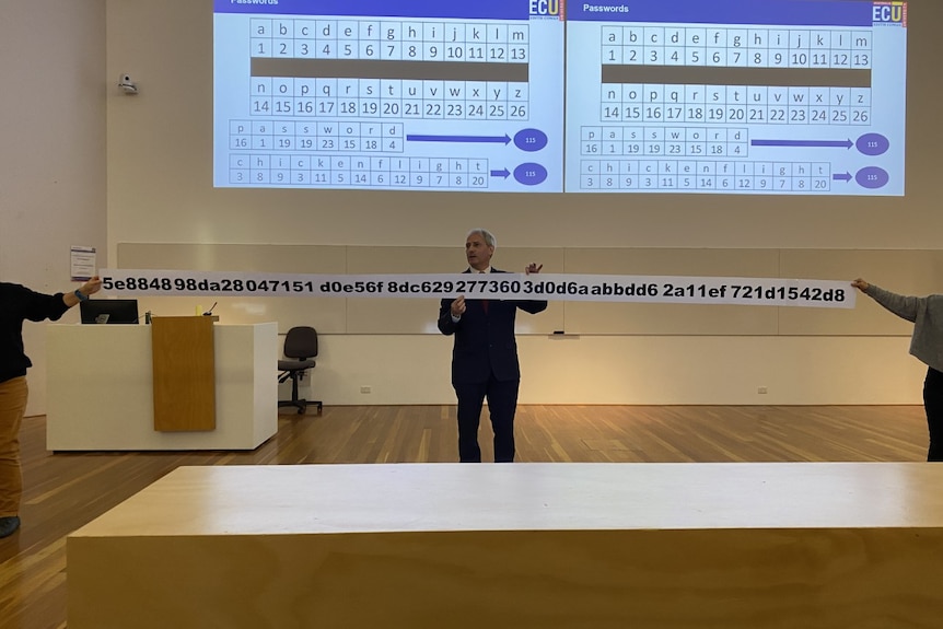 A man in a suit giving a lecture holds a ridiculously long piece of paper containing a password of random letters and numbers.