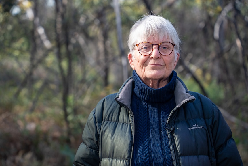 A woman standing in the bush, in warm clothing and glasses.