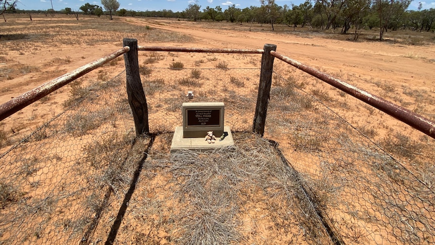 A fenced off grave marker surrounded by red sand and bushland.