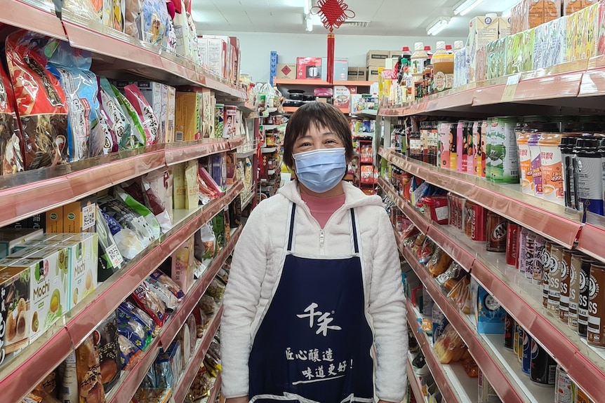 Jin Mei Chen, wearing a face mask and a blue apron, has smiley eyes as she stands in a grocery shop aisle.
