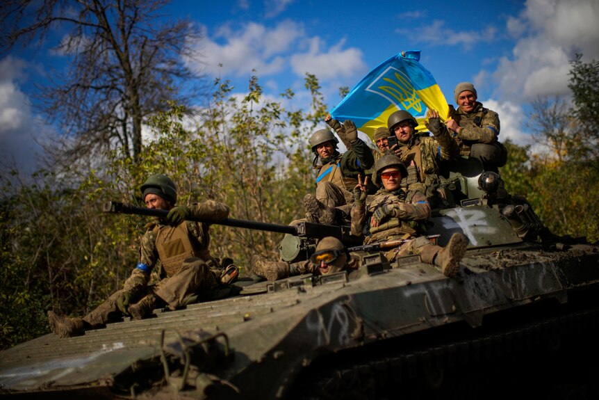 Ukrainian soldiers sit on an armoured vehicle holding a Ukraine flag.