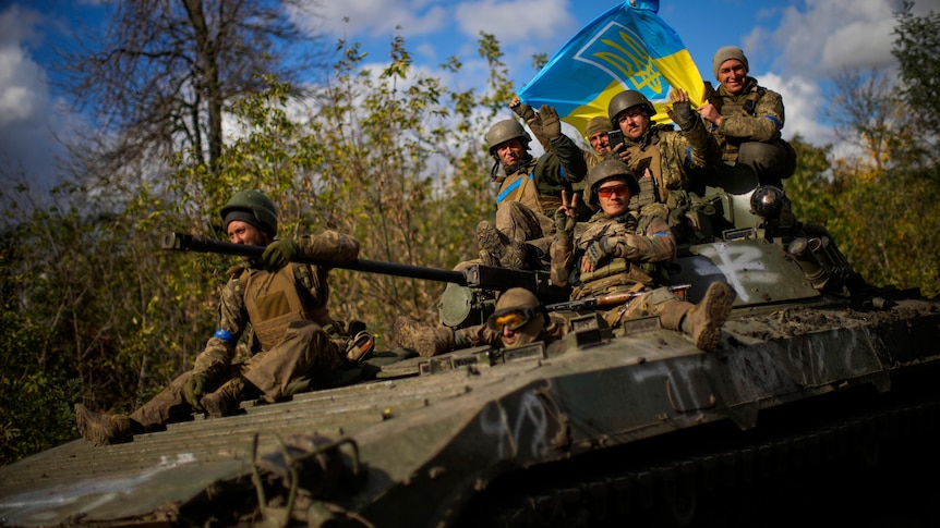 Ukrainian soldiers sit on an armoured vehicle holding a Ukraine flag.