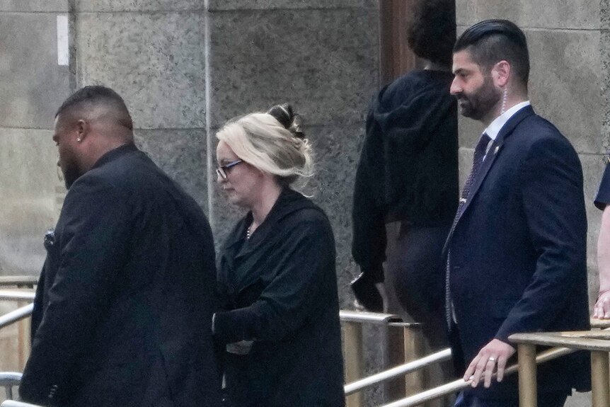 Stormy Daniels, dressed in black, walks out of a building, surrounded by men in uniforms.