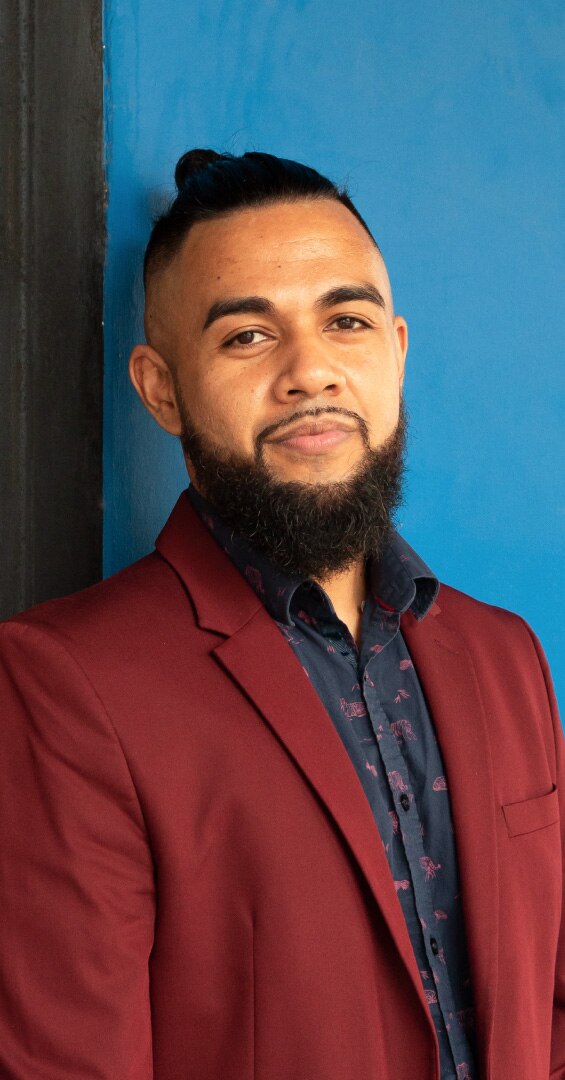 Man with black hair pulled into small bun, with beard and thin mustache, in red jacket and black shirt, smiling.