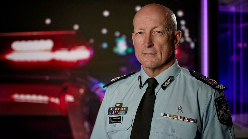 A bald man in a police uniform stands in front of a police background.