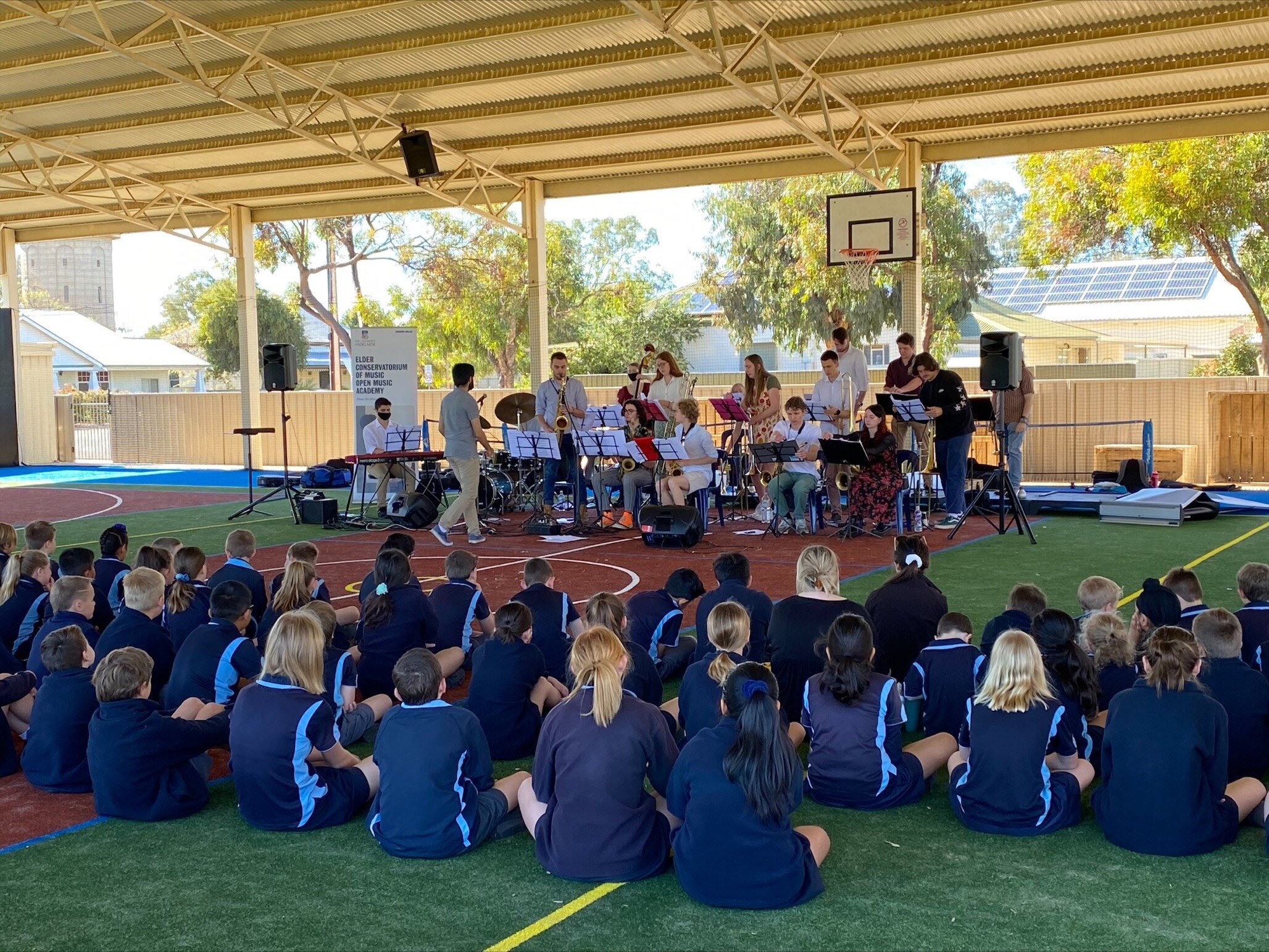 School students in blue uniform sitting on undercover court with band in front of them performing