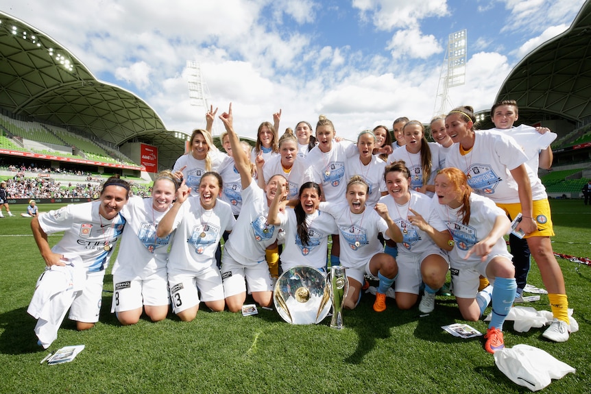 A soccer team wearing white and light blue pose on the field after winning two trophies