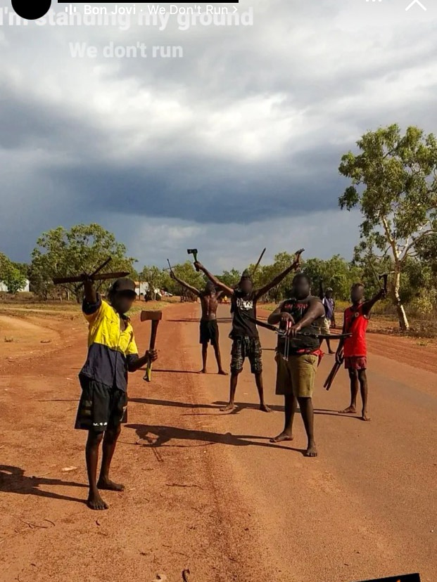 A group of men holding weapons including axes and crossbows, standing on a red dirt road in front of a remote community.