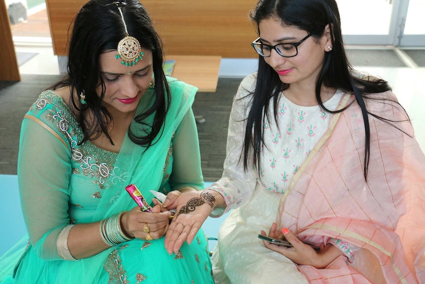 Two women sit indoors as one draws henna art on the other's skin.