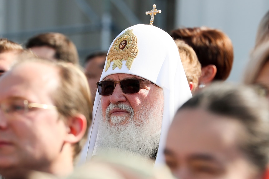 A man wearing a white headscarf embossed with a gold symbol and cross wears sunglasses in a crowd of people.