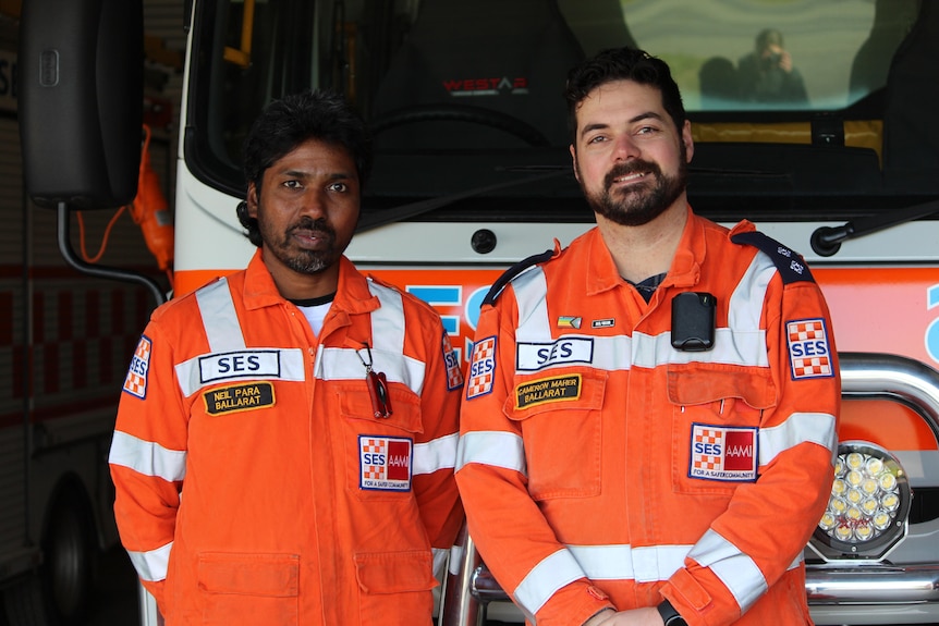 Two men stand in front of an SES truck in orange SES uniforms. 