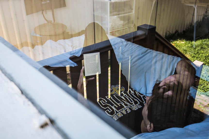 Colour photograph through a glass window of man reading an iPad in bed on a sunny day.