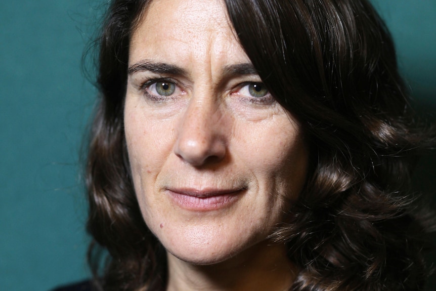 Esther Freud seen close up, with shoulder-length brown hair and dark jumper, against green background, smiling slightly.