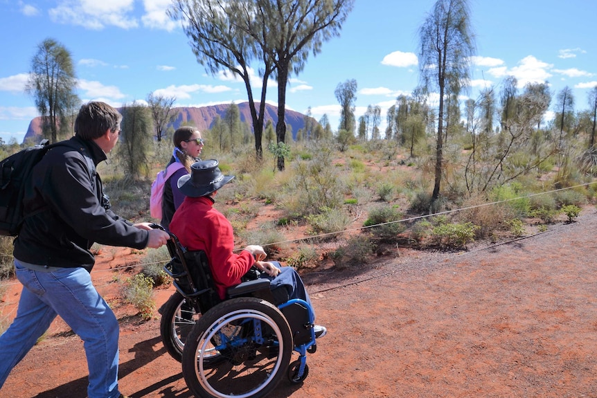 A man pushes a boy in a wheelchair while a girl walks alongside with Uluru in the background