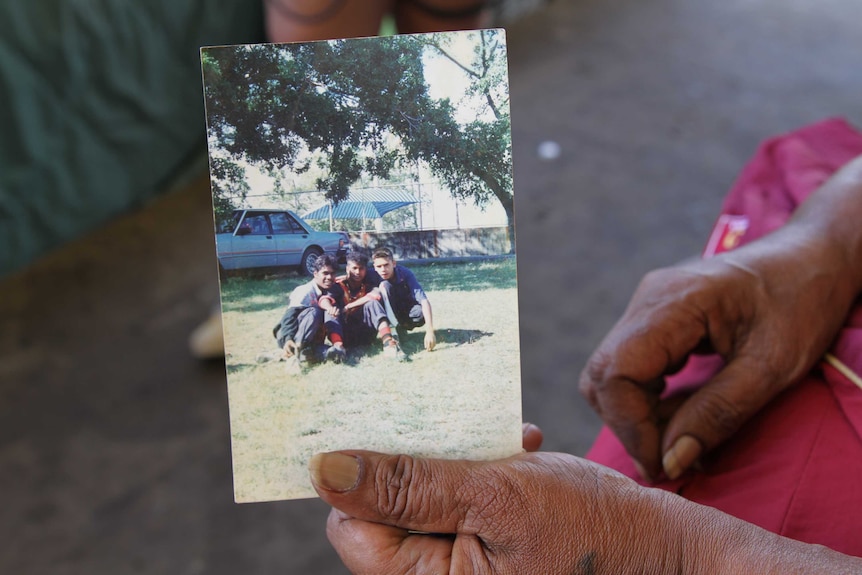 A hand holds a photo of Daniel Yock and two other teenage boys, sitting in a park.