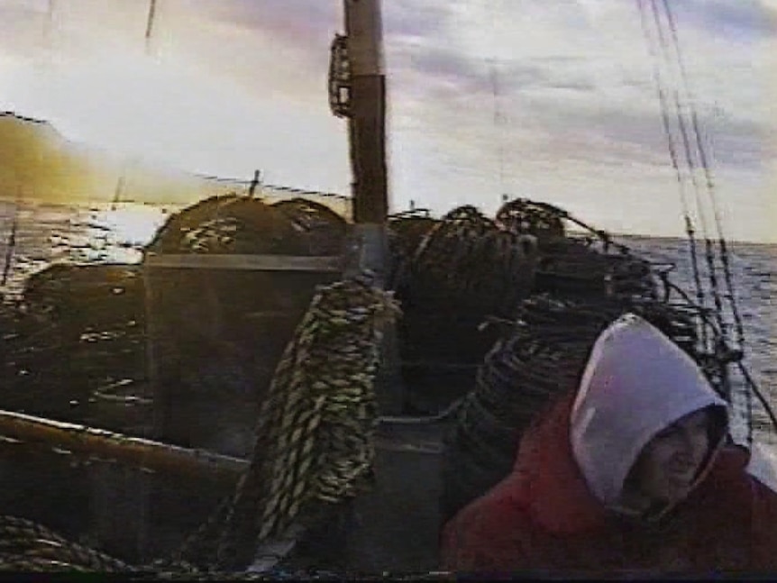 Deckhand in hoodie jacket rests on a craypot as crayboat cruises towards a sunrise.