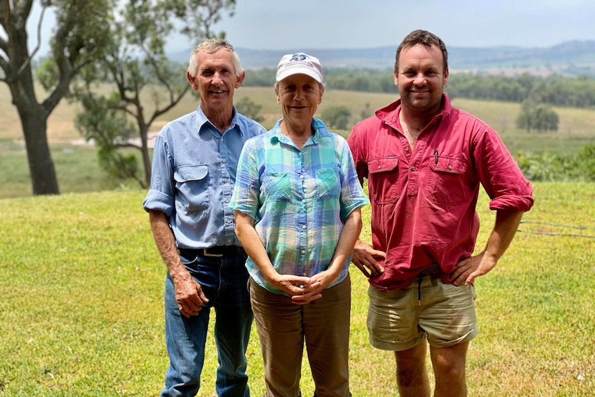 An elderly man and woman stand beside a younger man in a red shirt, with farmland behind them.