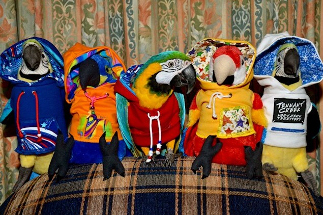 A parrot in a hoodie sits with other stuffed parrots in hoodies on a couch.