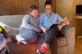 A mother and her pre-teen son sitting on a couch reading a book together.