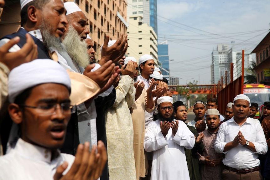 A group of men hold their hands out in a prayerful position, some appear to be singing.