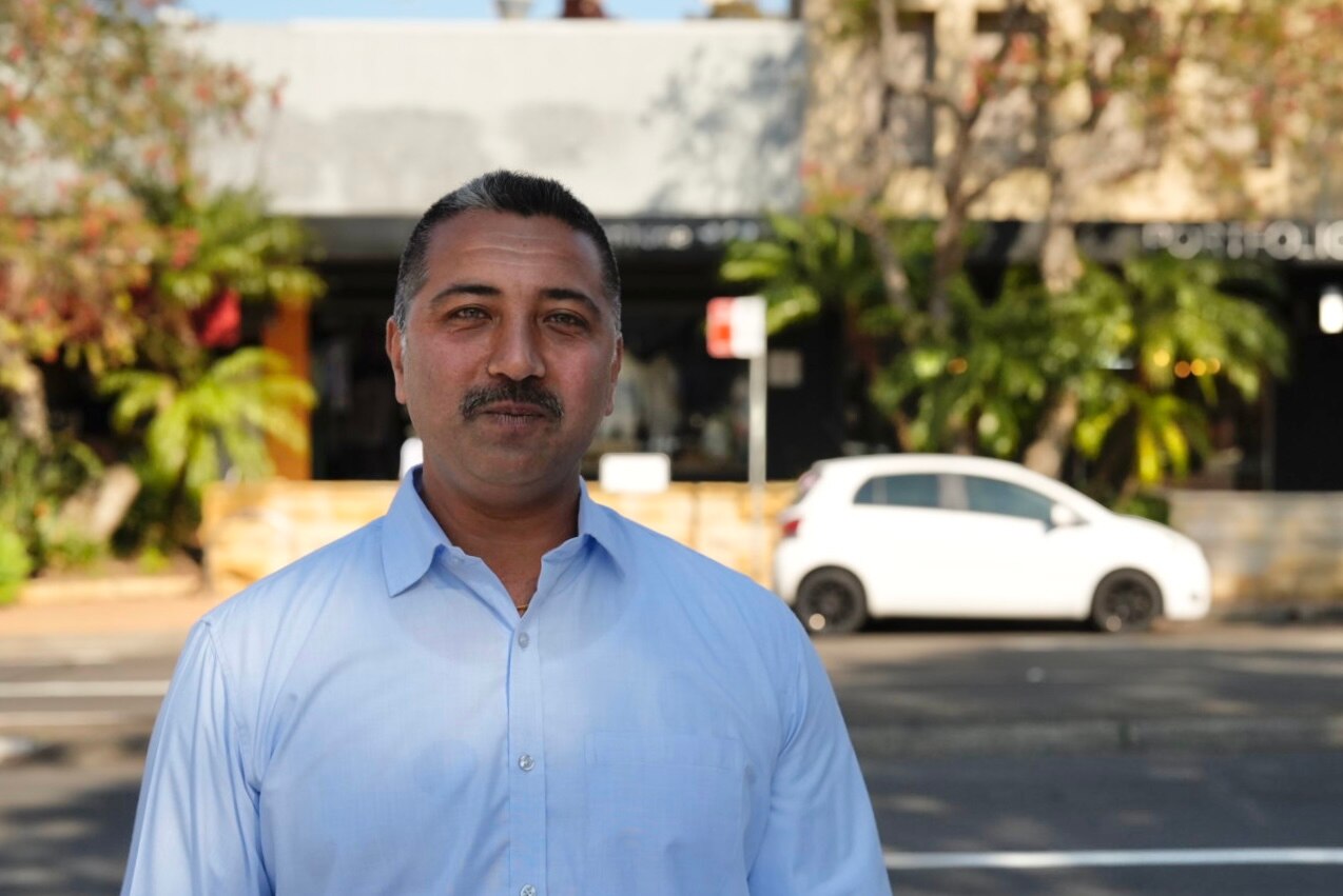A man wearing a white shirt smiles softly at the camera, standing on the street with a white car in the background