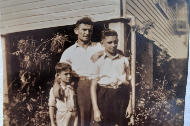 Old Black and white photo of a man standing behind two boys, in front of a suburban house, with a hand resting on each.