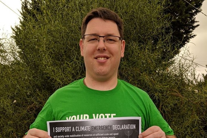 Jay Dessi holding a climate change poster while wearing a Greens t-shirt.