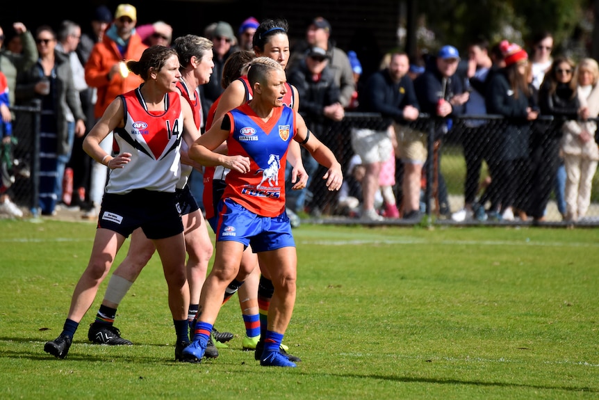 Two women Aussie Rules players stand, looking at the ball off camera. 
