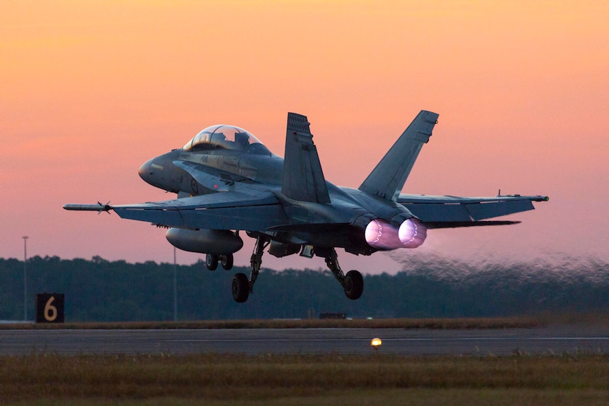 A fighter jet takes off away from the camera at dusk, with white-hot flames firing out of the the engine's afterburners.