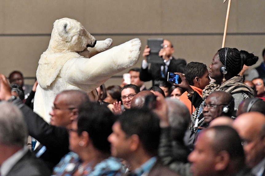A man in a polar bear costume walks between delegates at a conference.