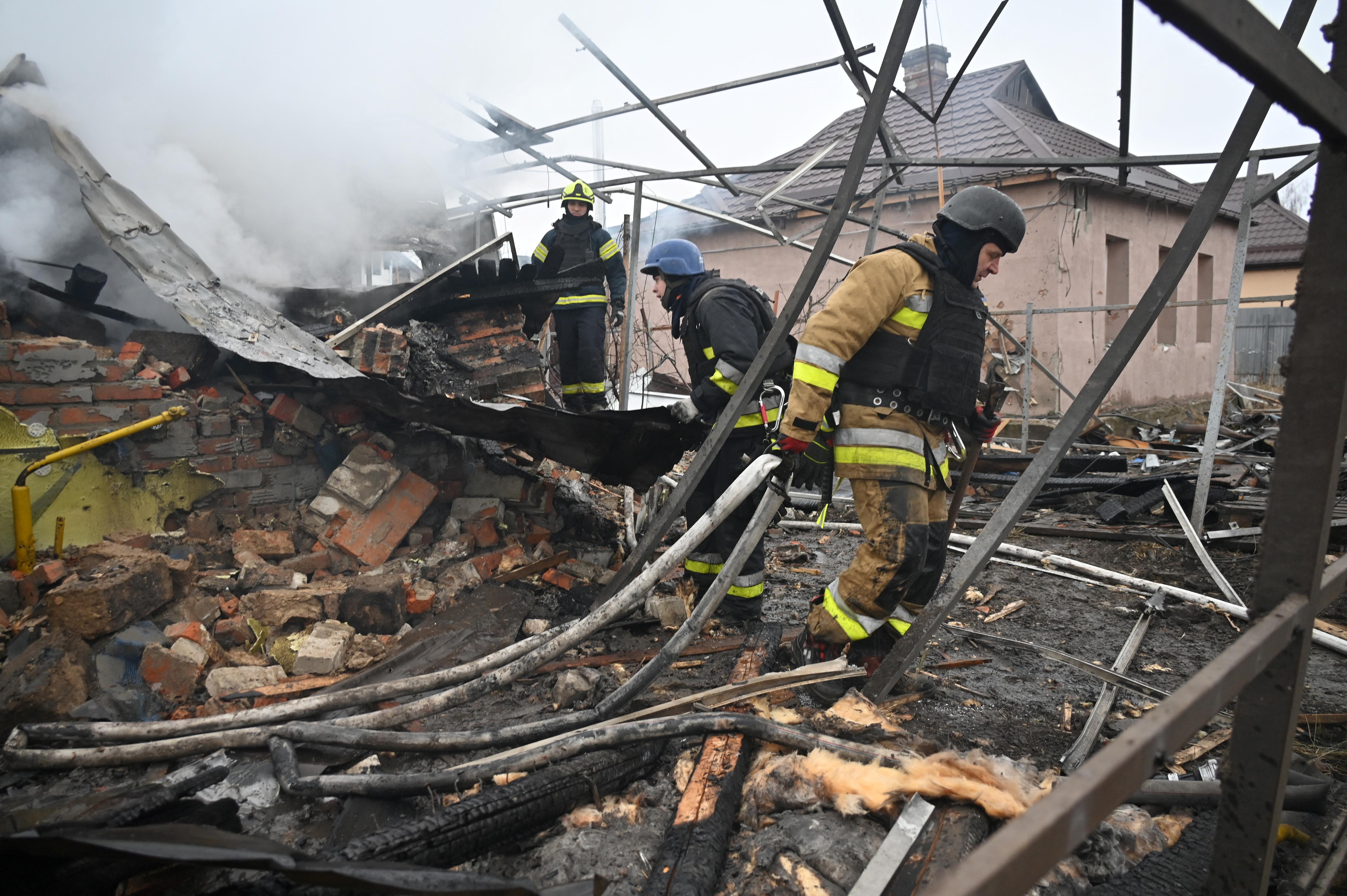 Three people stand amongst a pile of debris, with smoke rising behind them