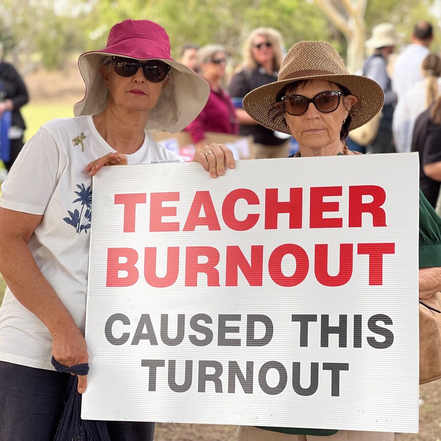Two female teachers stand outside during a rally, posing for a photo with a sign reading 'teacher burnout caused this turnout'.