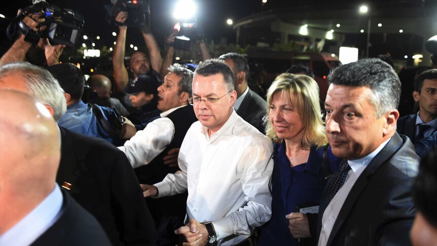 Pastor Andrew Brunson grasps the hand of his wife as they arrive at an airport surrounded by media.