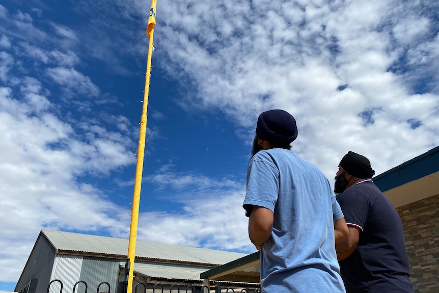 two men look up at a yellow flag pole, with a blue cloudy sky above them