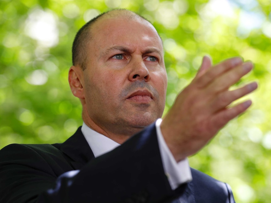 A white man wearing a suit pointing with his right arm and a frown expression with a blurred out tree behind him.