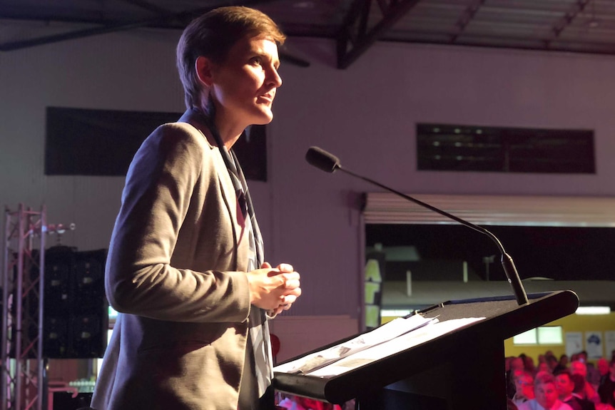 A woman with short, red hair addresses a room full of people from a podium. She is wearing a beige jacket