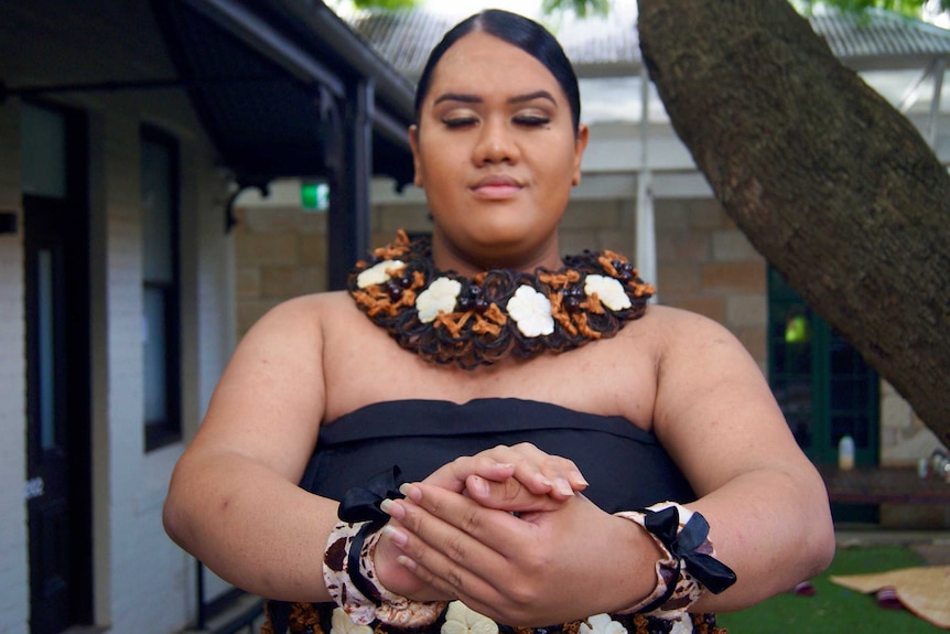 A transwoman wearing traditional Tongan cultural attire takes a peaceful pose with her eyes closed and her palms together.