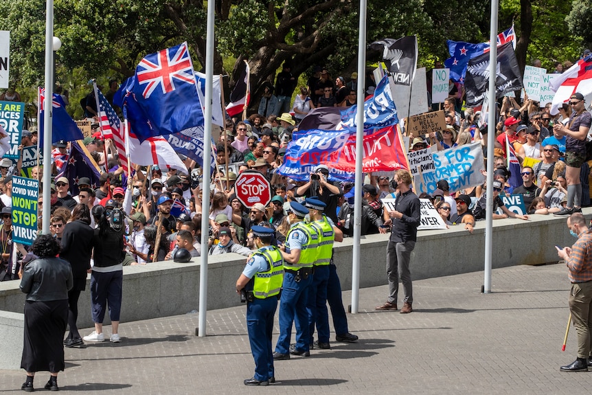 A crowd of people carrying placards and flags, including some that say TRUMP 2020, gather in an outdoor square. Police watch on