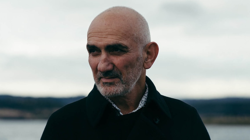 Photo of Paul Kelly standing outside wearing a dark jumper, looking past the camera. He has a bald head and a short beard.