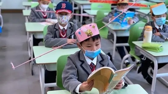 Chinese children sit at desks in neat rows in a classroom, all wearing hats with large wings sticking out the sides.