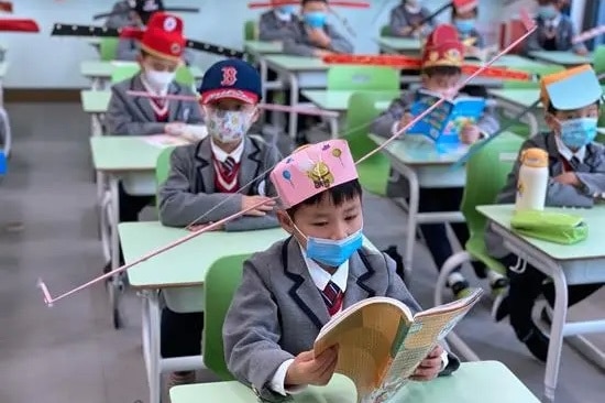 Chinese children sit at desks in neat rows in a classroom, all wearing hats with large wings sticking out the sides.