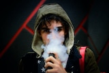 Young man with brown hair wearing hooded jacket while using a vape.