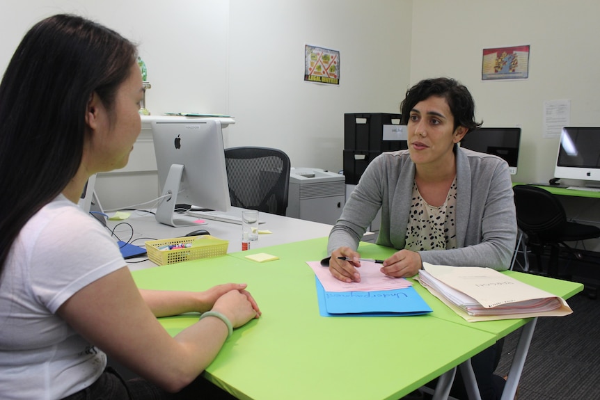 A woman sits at a desk and consults another woman.