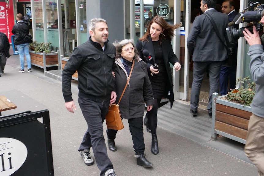 A woman with grey hair walks down the street arm in arm with a man as a reporter holds a microphone to her face.