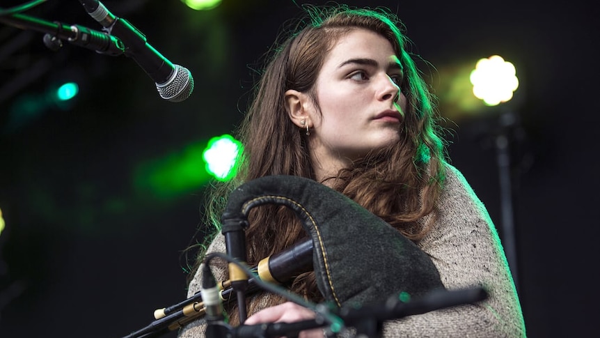 A woman is seated on stage performing and under her arm is a set of bagpipes.