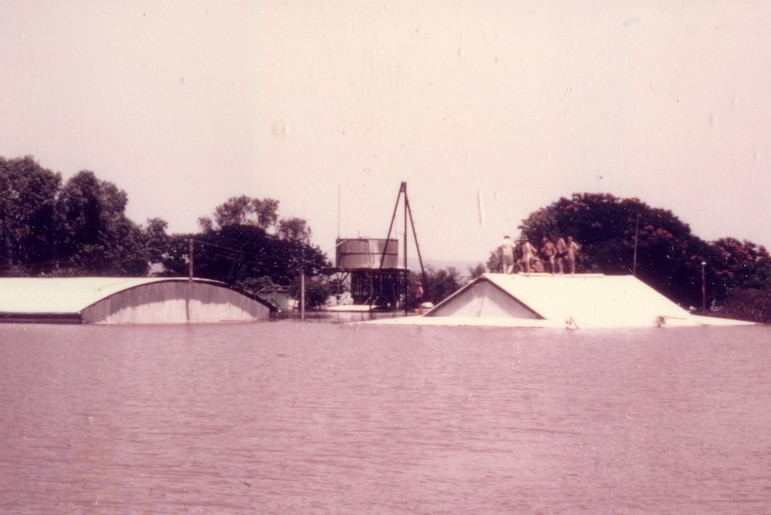 Five men stand on the roof of a building with floodwater up to the eaves.