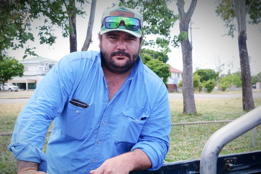 A man in blue shirt, hat and sunglasses, leaning over the edge of a ute tray. 