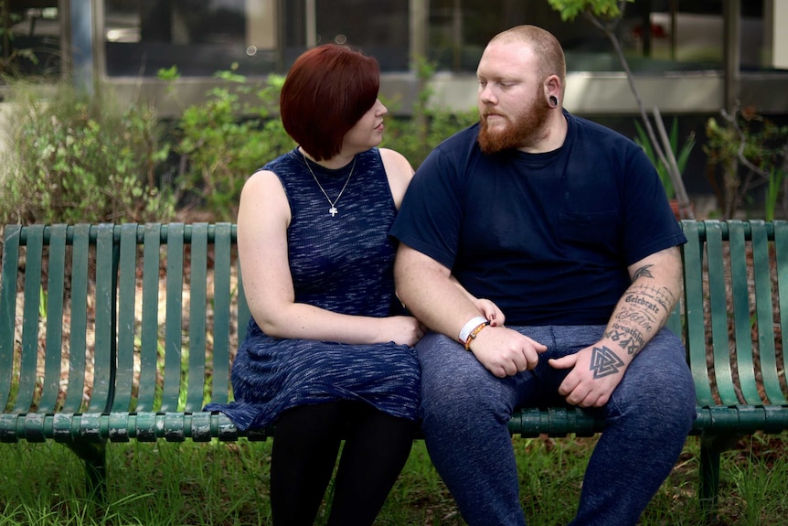 A couple sits on a park bench, looking at each other with love and concern.