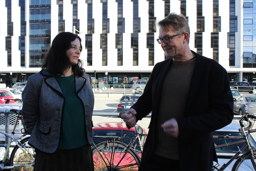 A woman and man in conversation on a street beside a bicycle rack.